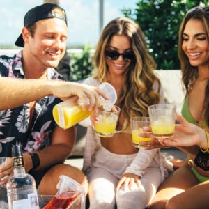 Male pouring juice into the glasses of two females while sitting in a poolside cabana